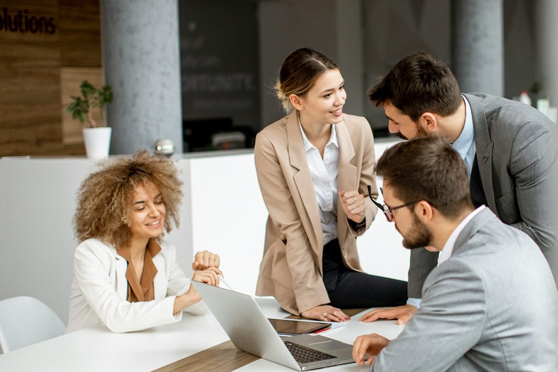 Business English Vier Personen in Geschäftsanzügen diskutieren an einem Tisch mit Laptops und Unterlagen.