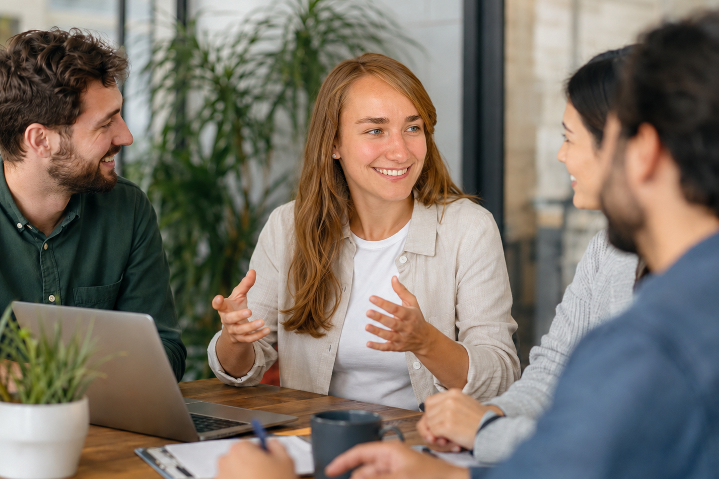 Business English Vier Personen in Geschäftsanzügen diskutieren an einem Tisch mit Laptops und Unterlagen.